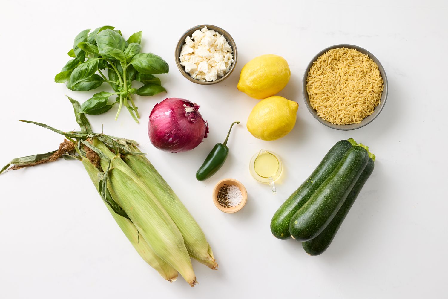Ingredients including corn zucchini lemon basil red onion feta cheese orzo jalapeo and seasonings arranged on a white surface