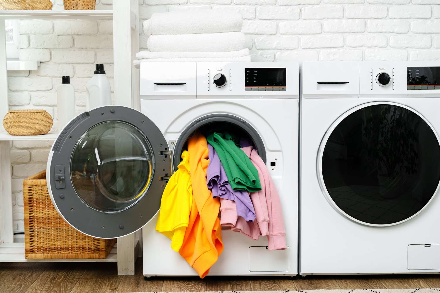 Frontloading washing machine with colorful clothes spilling out next to a matching dryer in a laundry room setup
