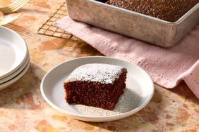 A slice of chocolate cake dusted with powdered sugar on a plate with a pan of cake in the background