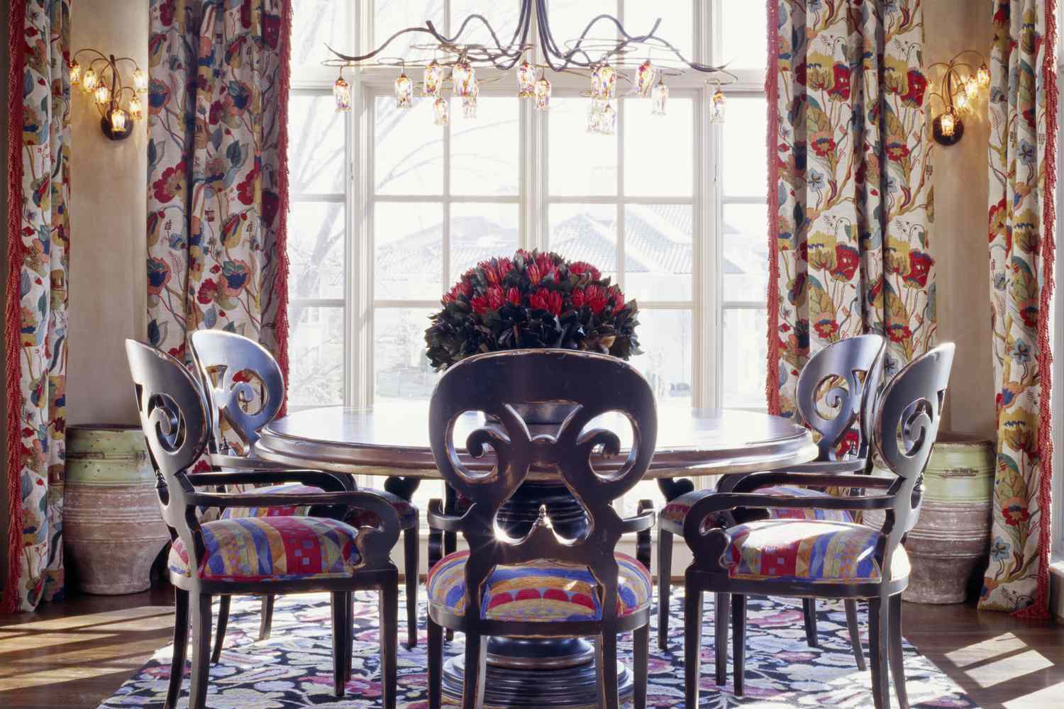 A dining room with a round table six ornate chairs floral curtains and a central window letting in natural light