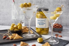 Two martini glasses garnished with olives a jar of blue cheese stuffed olives crackers almonds and a drink in the background