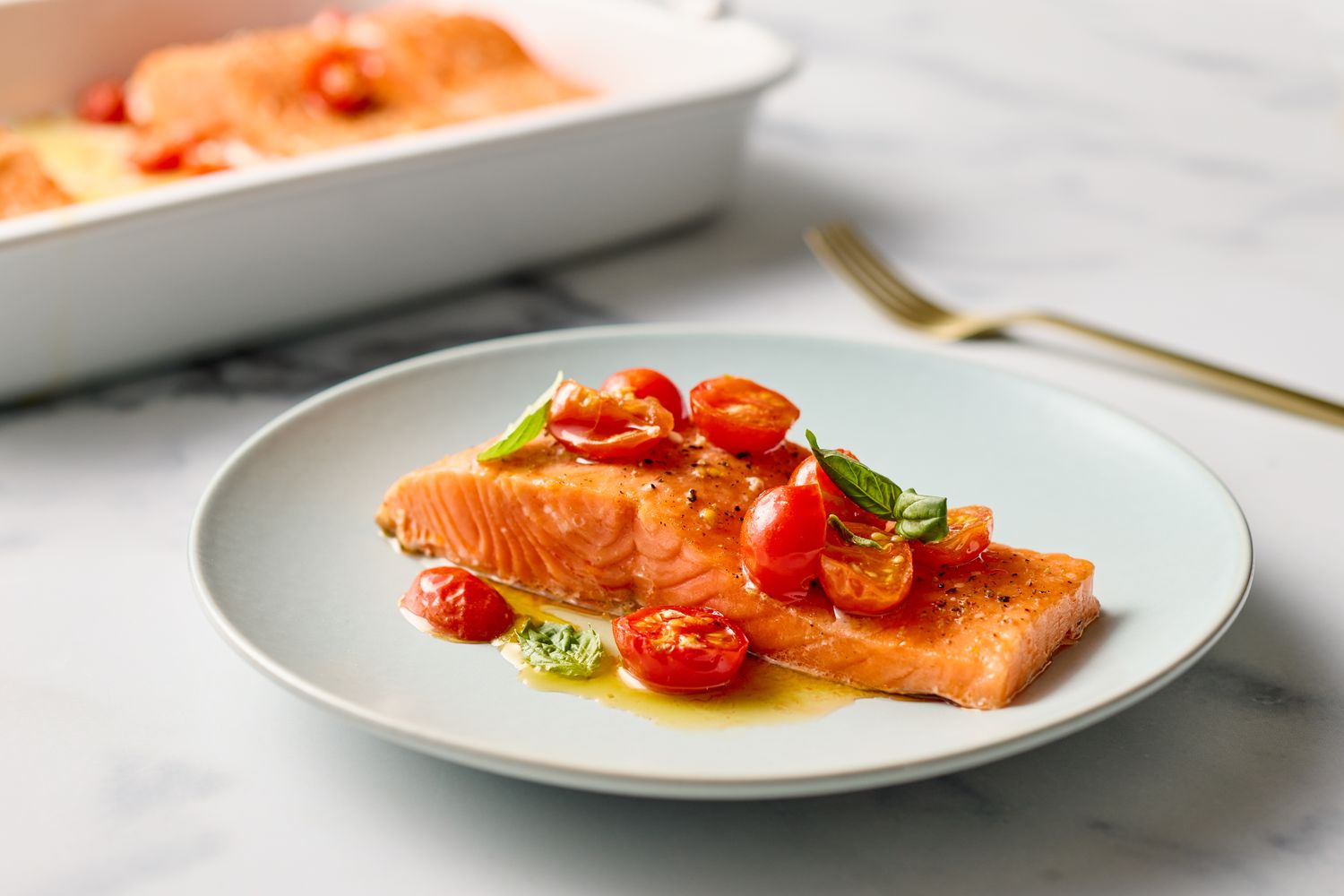 A plate with a portion of baked salmon garnished with cherry tomatoes and herbs a baking dish in the background