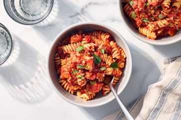 A bowl of pasta with tomatobased sauce and garnish served with water glasses and placed on a table with a napkin