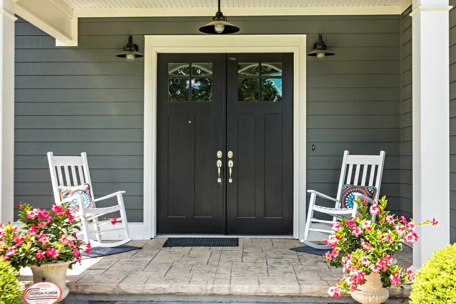 A front porch with a double door two white rocking chairs and flowering plants in decorative pots