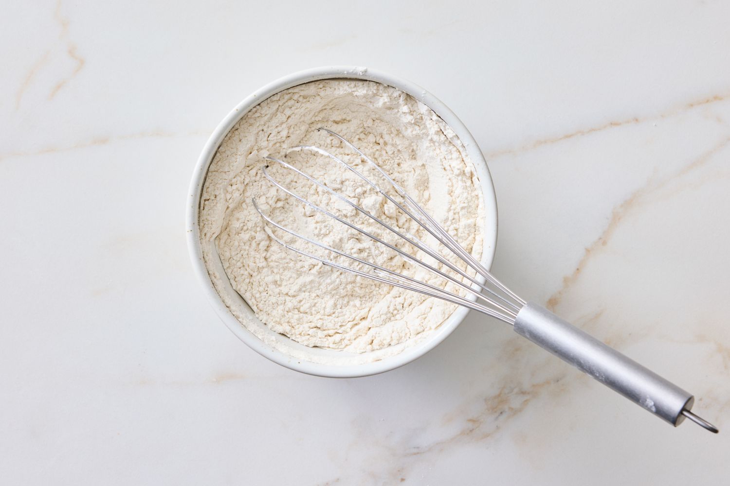 A mixing bowl with flour and a whisk on a countertop