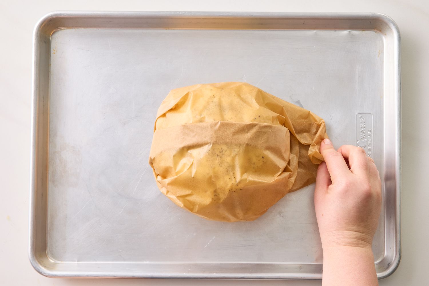 A hand wraps parchment paper around a head of cauliflower on a baking sheet