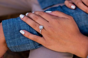 A hand wearing an engagement ring rests on a denimclad leg