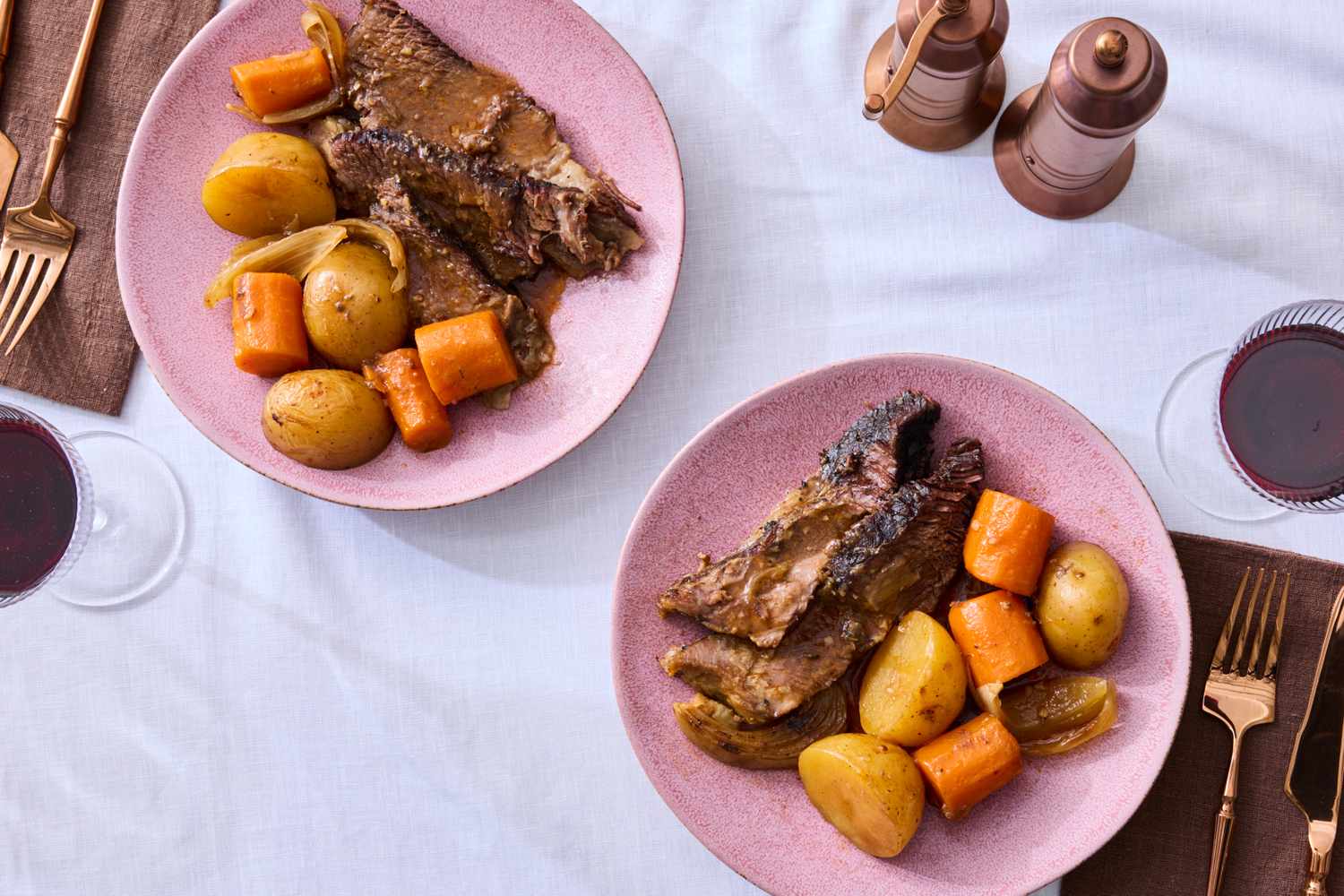 Two plates of pot roast served with potatoes and carrots accompanied by glasses of red wine and salt and pepper shakers