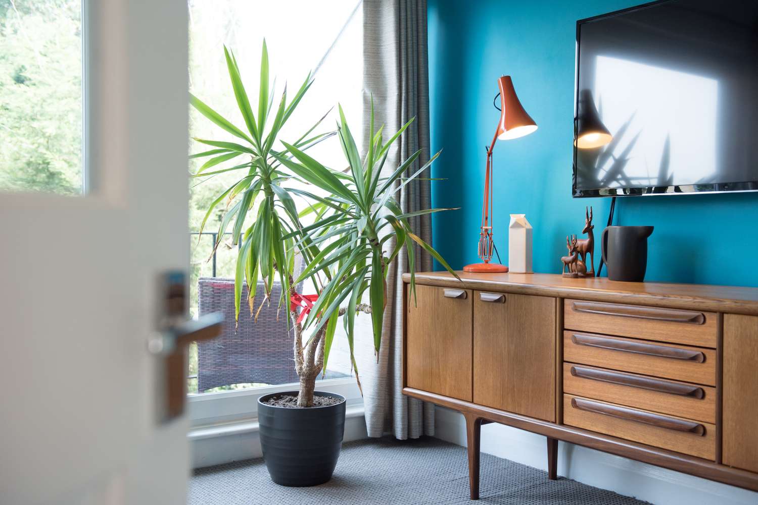 Midcentury modern living room with a wooden sideboard a potted plant a wallmounted TV and a lamp on the counter
