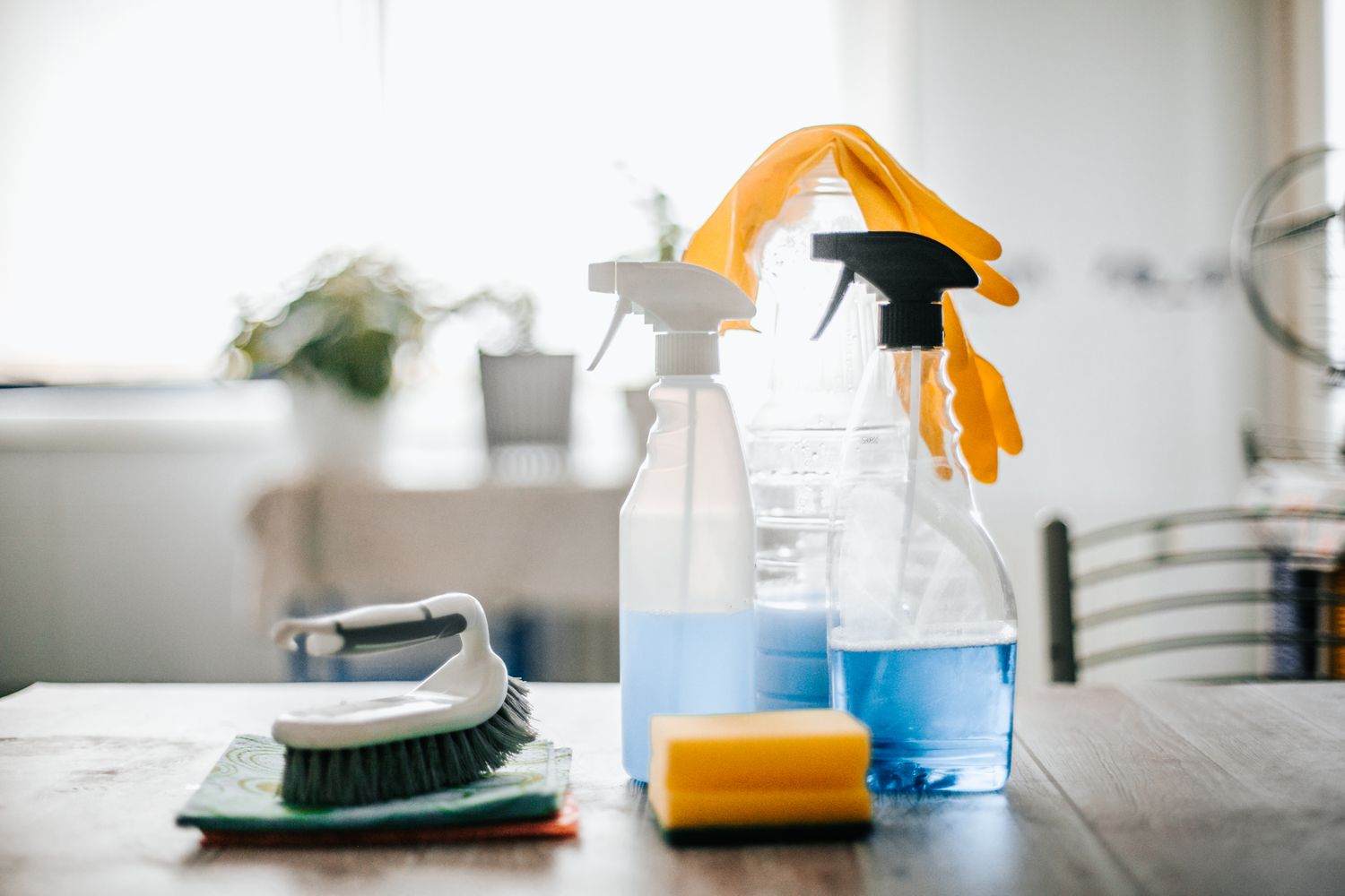 Cleaning supplies on a wooden table including spray bottles sponges a scrub brush and rubber gloves