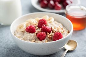 A bowl of oatmeal topped with raspberries banana slices and chopped nuts with honey and cherries in the background