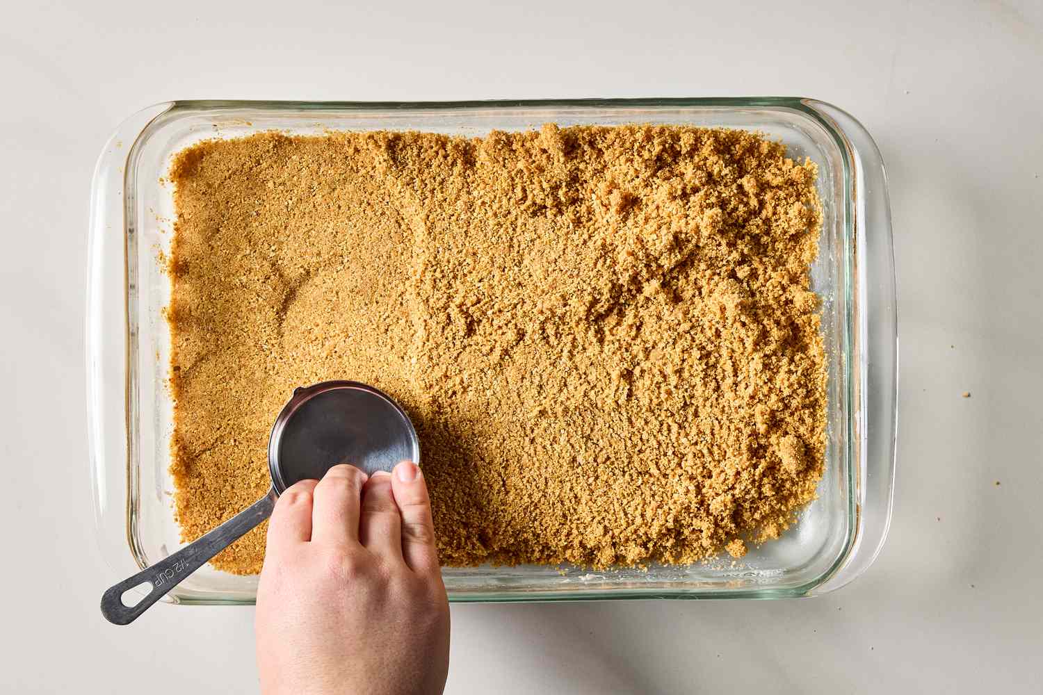 A hand compressing a graham cracker crust into a baking dish with a measuring cup