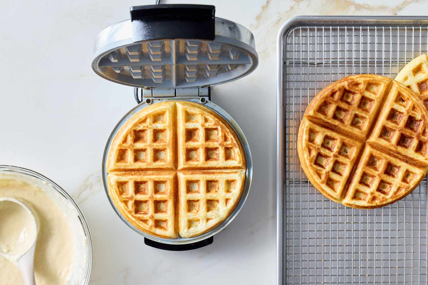 Waffle maker with a cooked waffle batter in a bowl and another waffle on a cooling rack