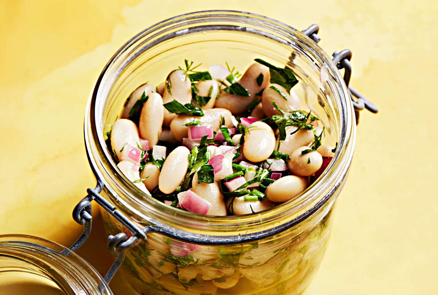 A jar filled with bean salad including white beans chopped herbs and diced red onions