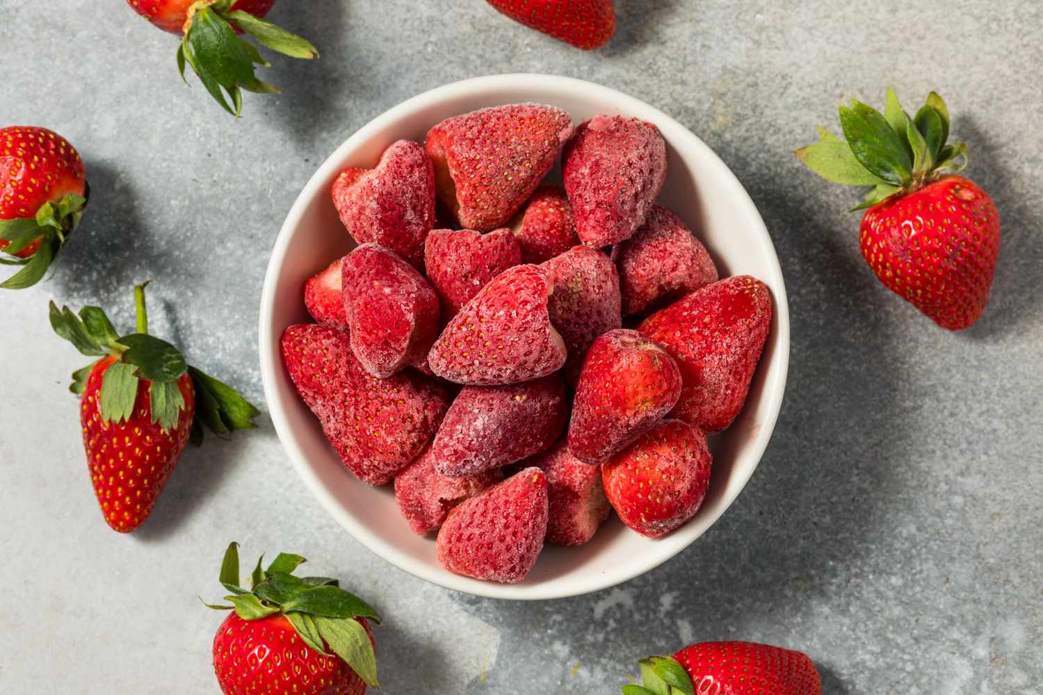 A bowl of frozen strawberries with fresh strawberries around it on a gray surface
