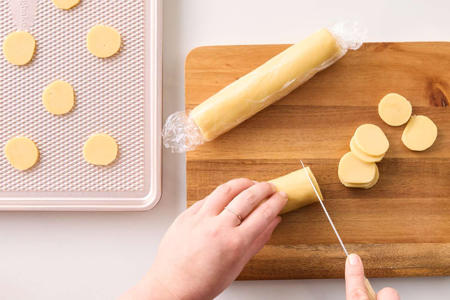 Hands slicing dough into discs on a cutting board uncooked cookies arranged on a nearby tray