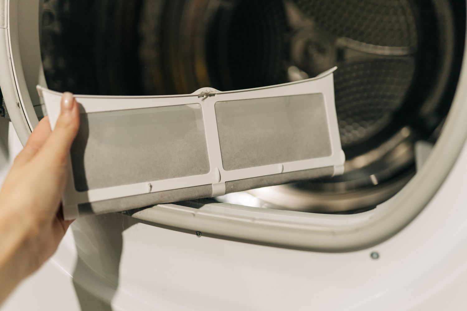 A hand holding a dryer lint filter in front of an open dryer drum