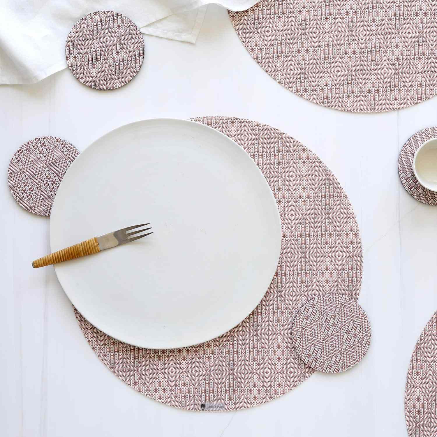 A round plate on a patterned placemat with a fork placed on it surrounded by a cup and coasters