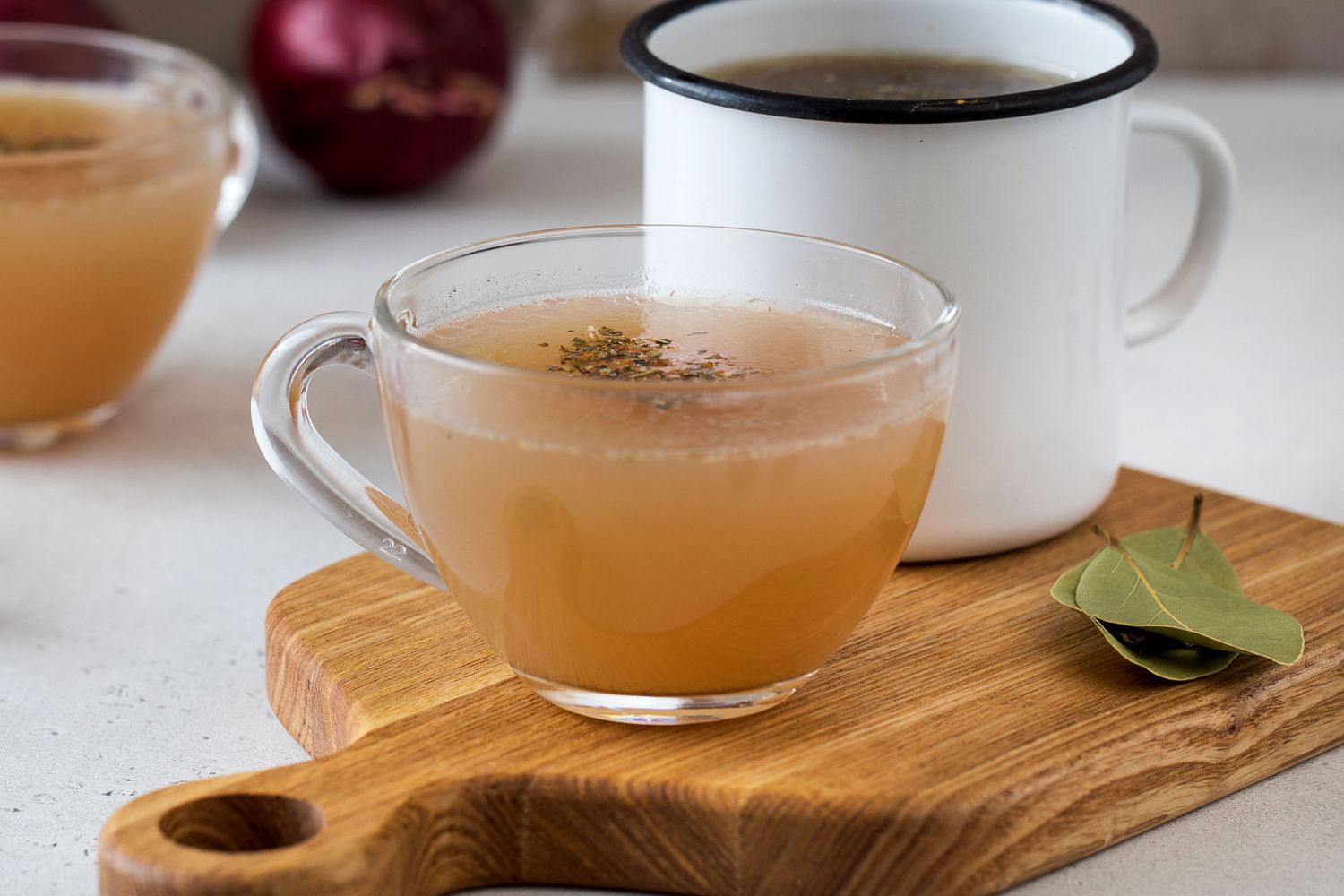 A glass mug of broth on a wooden board with a bay leaf next to an enamel cup in the background