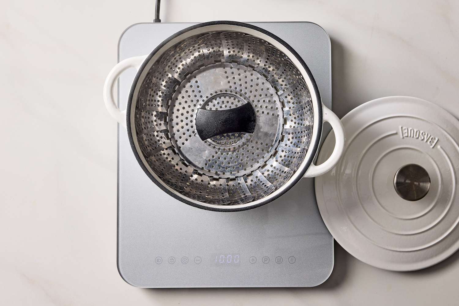 Metal pot containing a steamer basket placed on an induction cooktop with a lid to the side