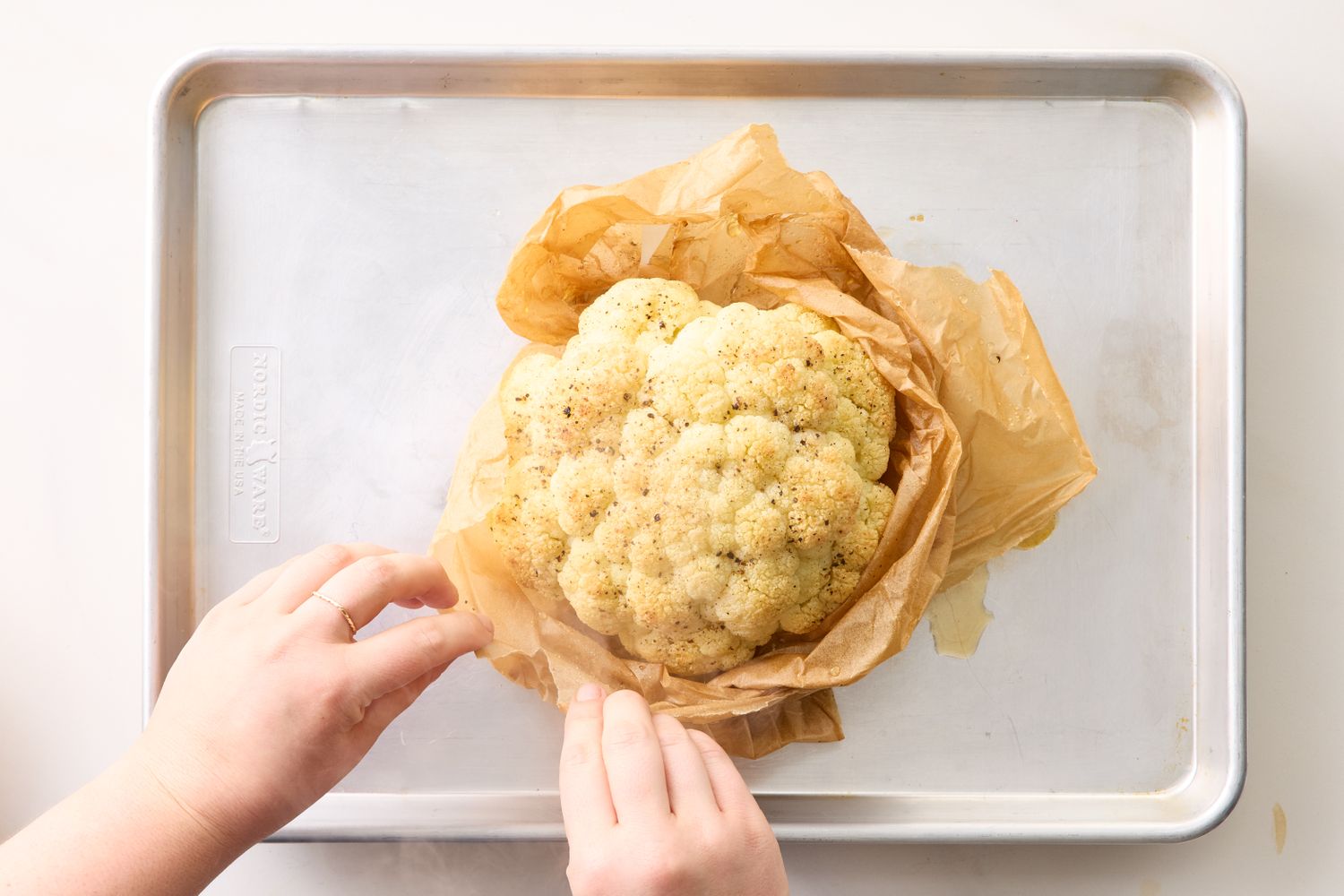 Whole roasted cauliflower on a baking sheet being wrapped in parchment paper by hands