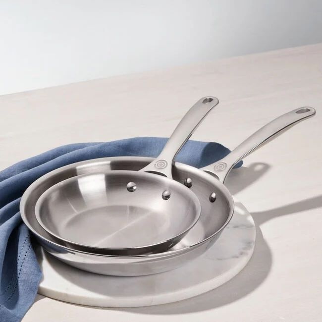 Two stainless steel frying pans stacked placed on a light countertop next to a cloth