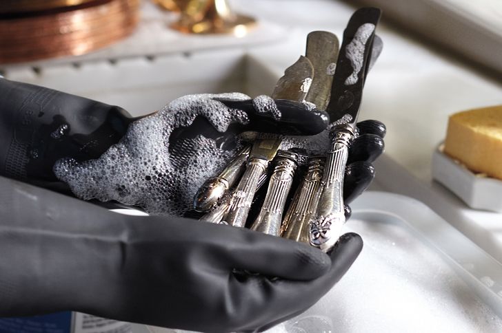 Hands wearing gloves cleaning silver utensils with soap suds