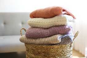 A bunch of stacked knitted pastel color sweaters with different knitting patterns perfectly folded in wicker basket on table, living room background.