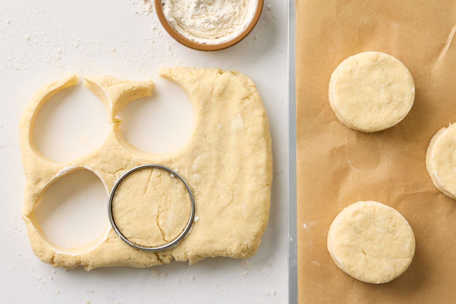 Dough with circular pieces cut out and a metal cutter nearby alongside parchment paper with uncooked rounds of dough