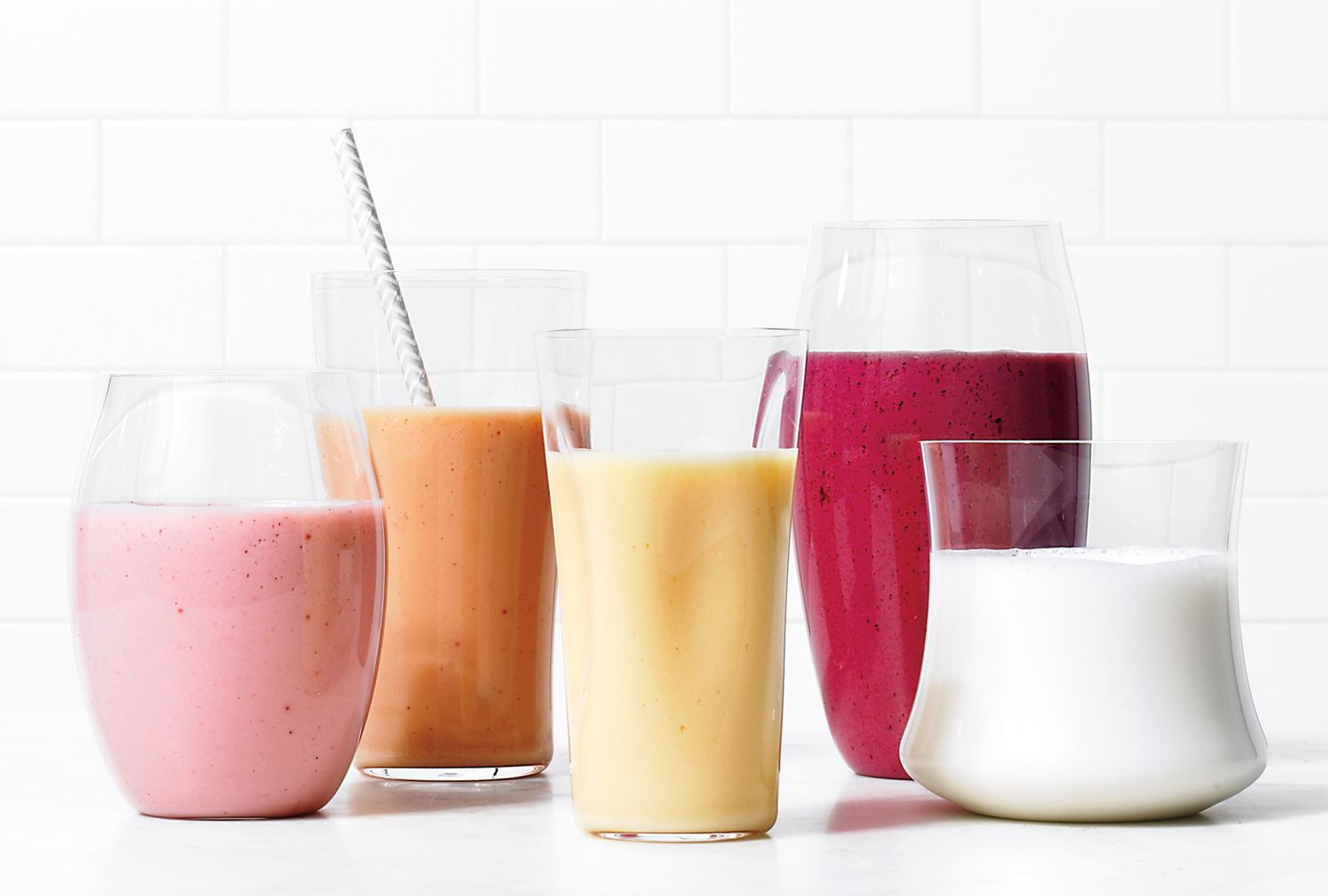 An assortment of smoothies in various glasses displayed on a white tile background