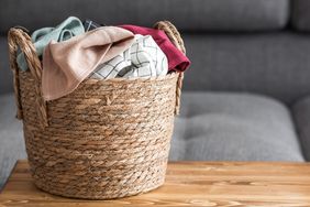 A woven basket containing various clothes placed on a wooden surface with a couch in the background