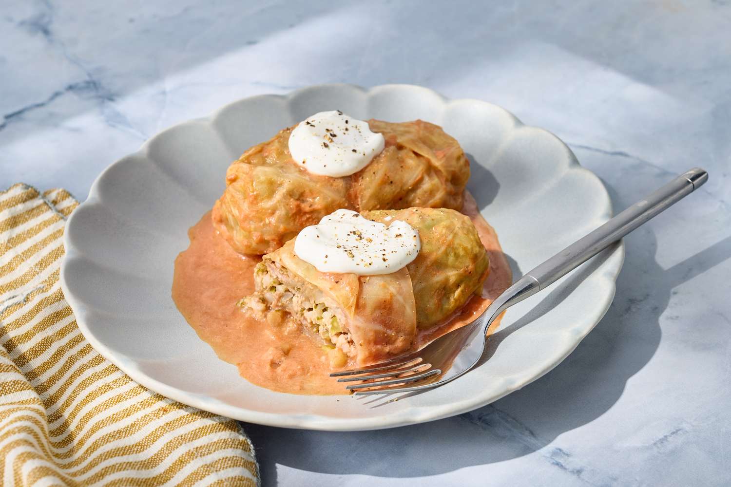 Two stuffed cabbage rolls on a plate with sauce and dollops of cream accompanied by a fork on a striped napkin