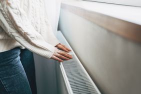 A person wearing a knitted sweater and jeans touching a radiator with their hands in an indoor setting