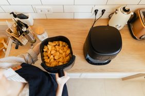 A person holds a basket of cooked potato pieces next to an air fryer on a wooden kitchen counter