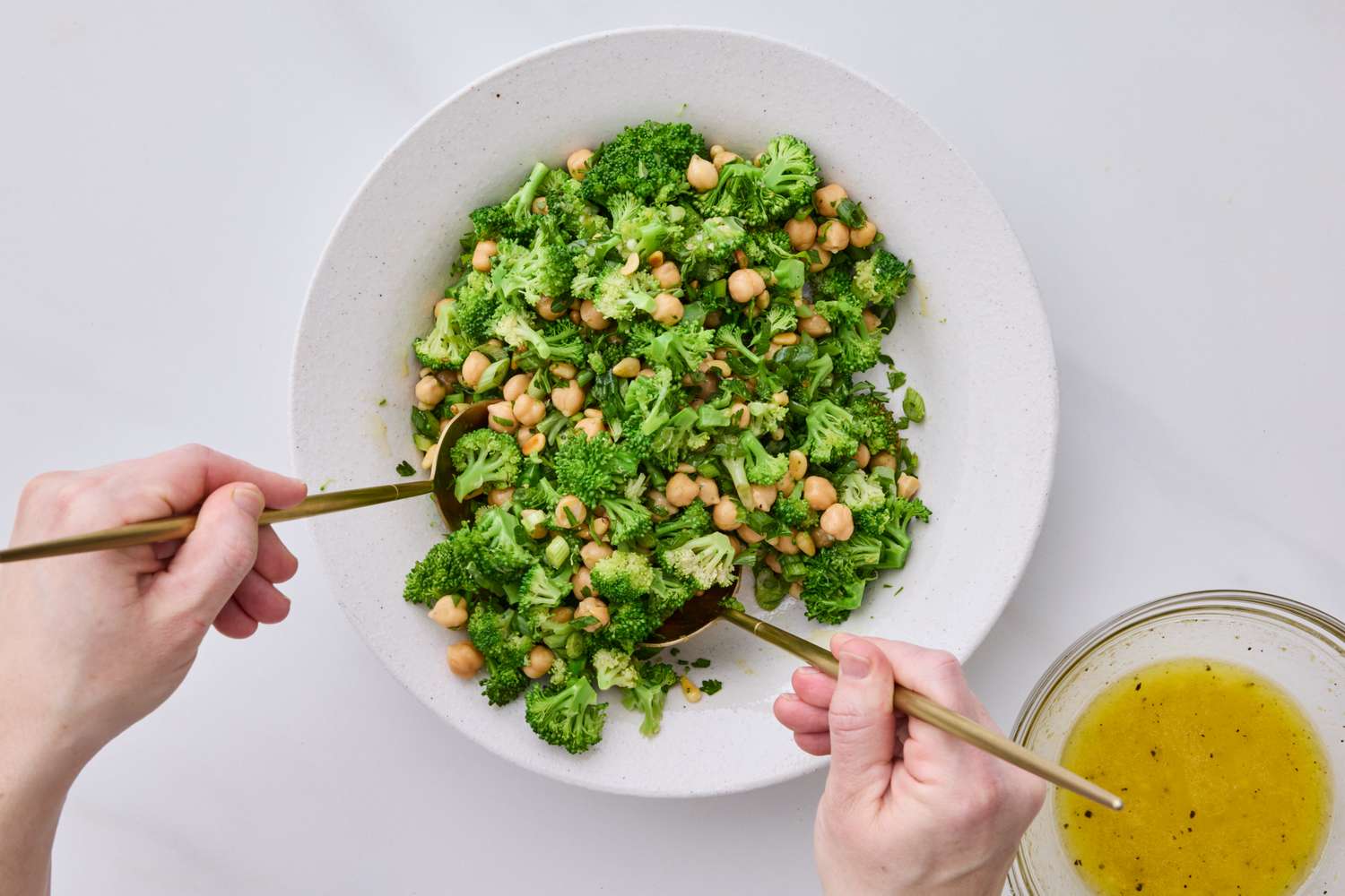 A person tossing broccoli and chickpea salad in a bowl with dressing on the side