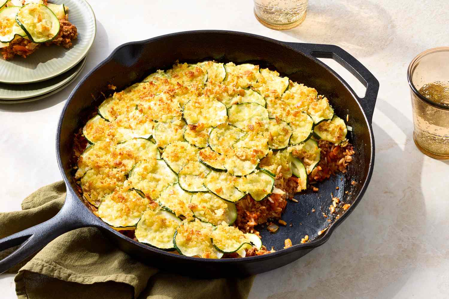 A skillet filled with a beef and rice casserole topped with thinly sliced zucchini and a breadcrumb layer set on a table
