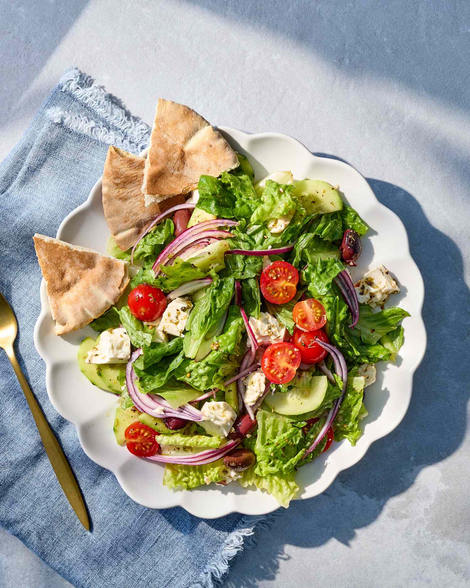 A plate of Greek salad with lettuce, cherry tomatoes, feta cheese, red onion, cucumber, and pita bread wedges