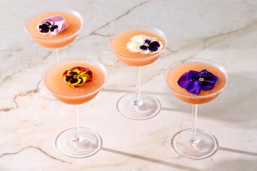 Four cocktails in stemmed glasses garnished with edible flowers arranged on a marble surface