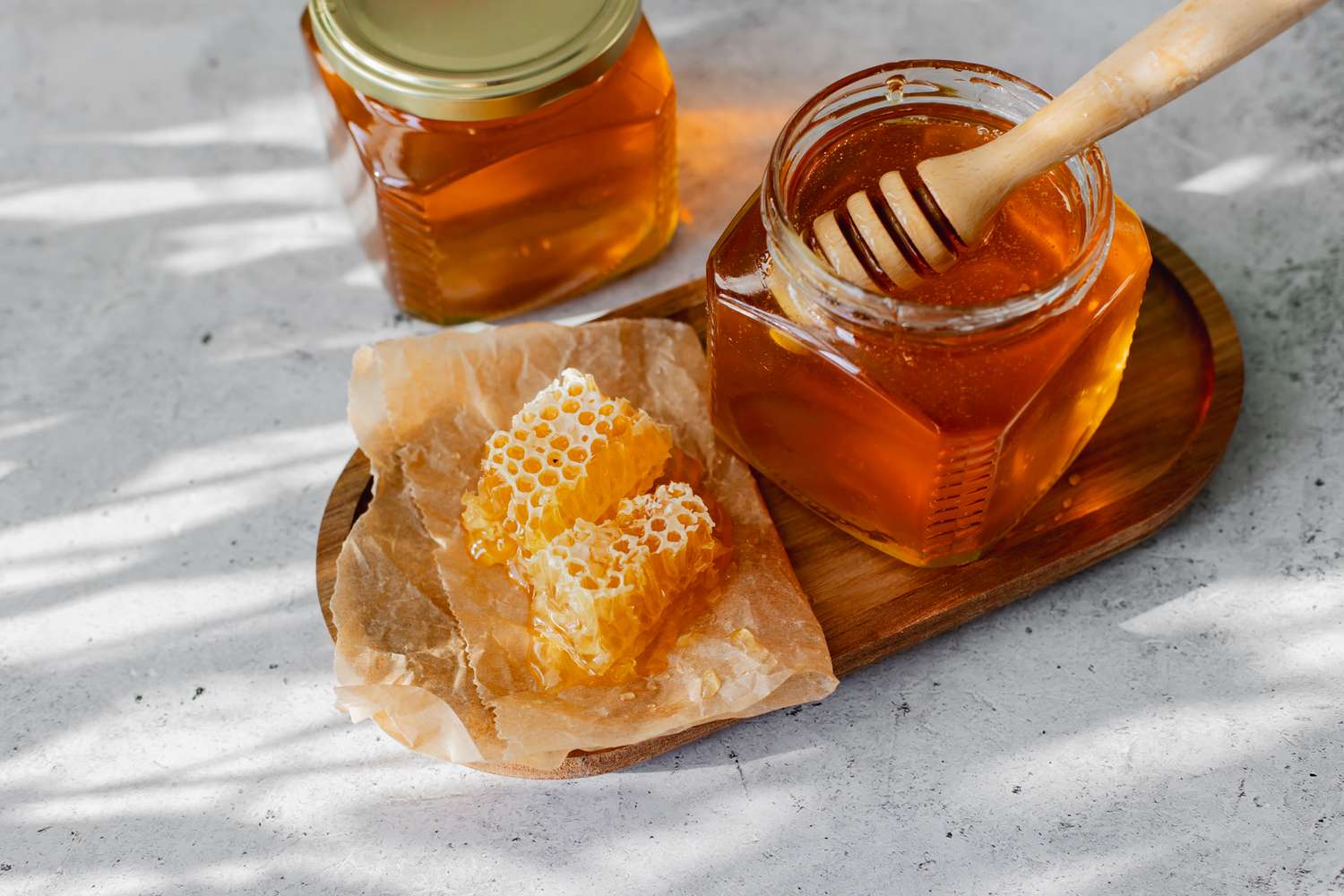 honey in two glass jars, one with lid off and honey dipper, also honeycomb on paper in dappled light