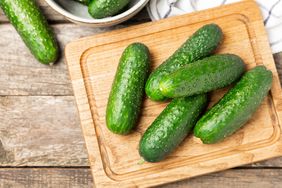 A wooden cutting board with several cucumbers placed on it additional cucumbers in a bowl in the background table surface underneath