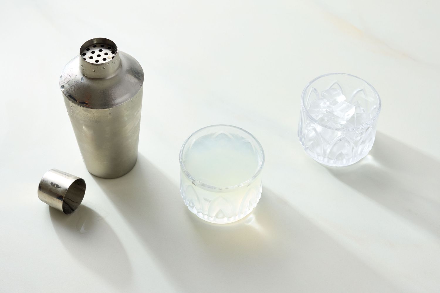 Cocktail shaker a glass with a clear beverage and a glass with ice cubes positioned on a light surface