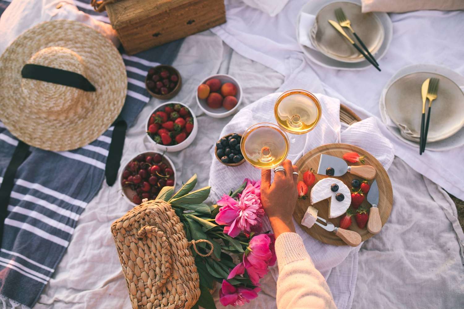 Picnic setup with food and drinks on a blanket outdoors featuring two glasses of a beverage being held up in a toast