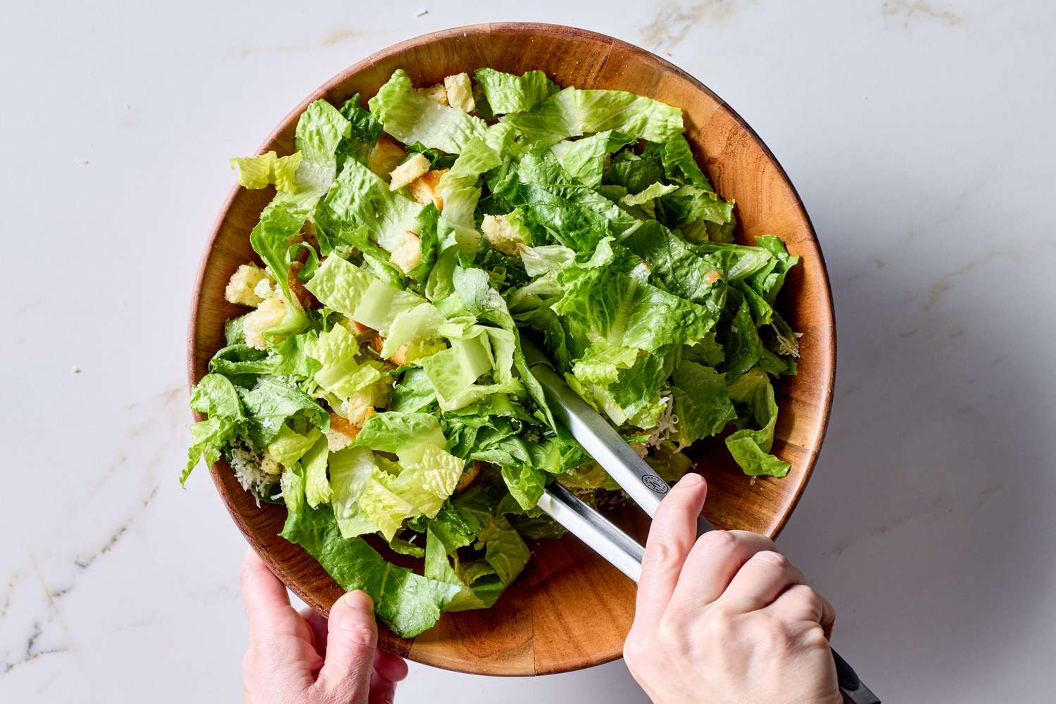 Someone mixing a bowl of Caesar salad with tongs