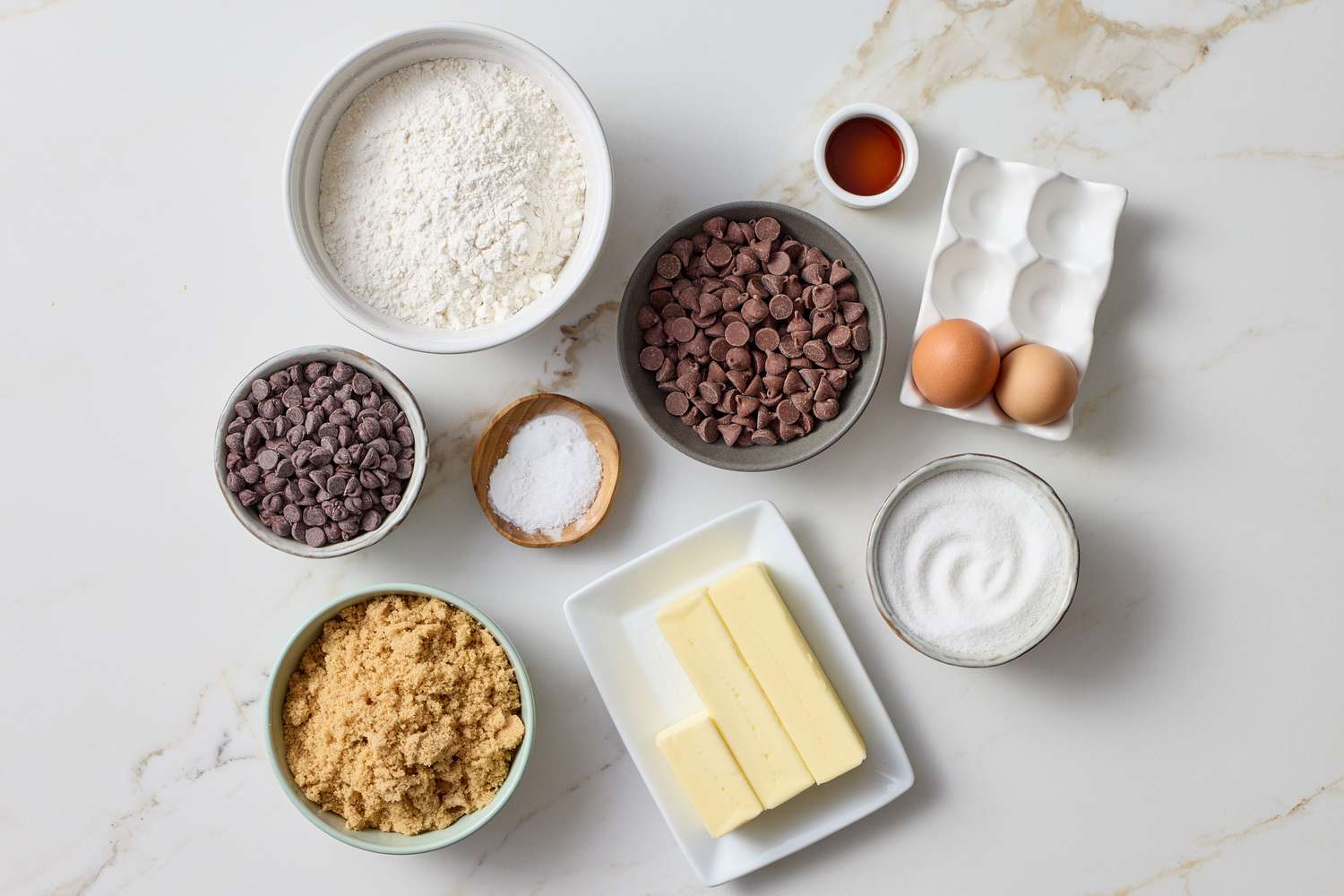 Ingredients for making chocolate chip cookies arranged on a surface including flour sugar eggs butter chocolate chips and vanilla extract