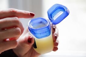 A hand opening a container of petroleum jelly with a blue lid another finger reaching to touch the contents