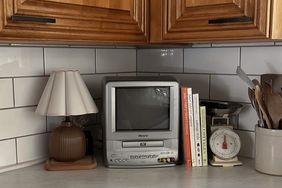 A small television sits on a kitchen counter surrounded by various items including books a lamp and a kitchen scale