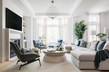 Minimalist living room by Outline Interiors: beige sectional sofa, blue accent chairs, wood coffee table with books, stone fireplace, lush potted plants, TV, and large windows with sheer curtains. Light wood floor and natural light create a calm, stylish space.