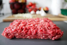 A block of raw ground beef on a cutting surface with food preparation items in the background