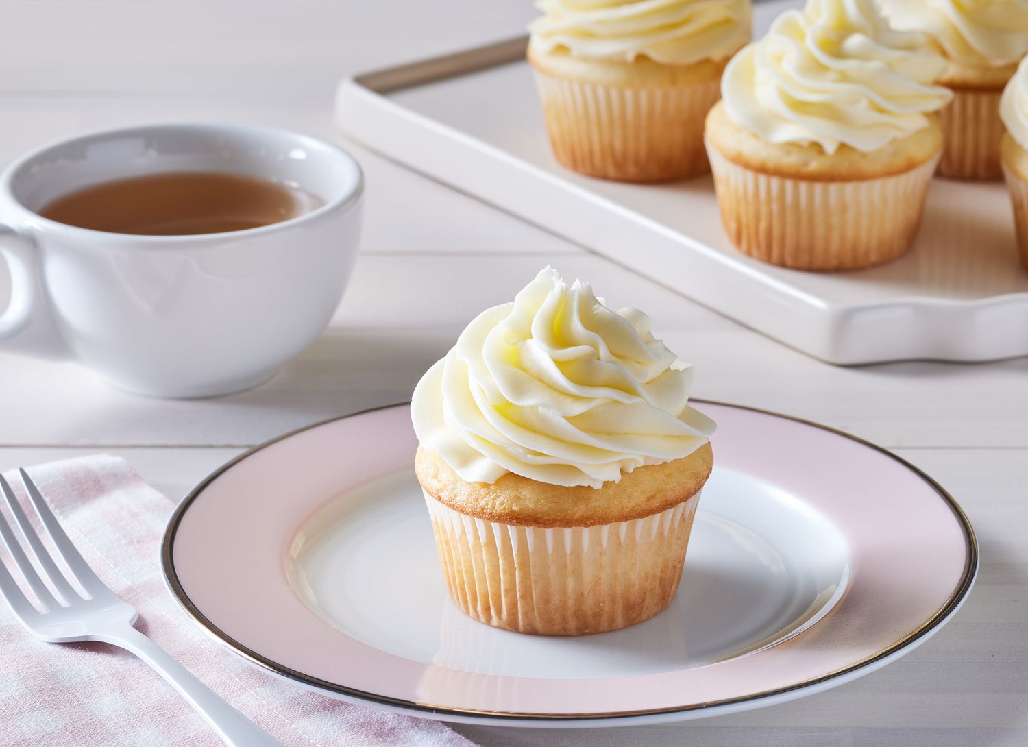 pale pink plate with cupcake with white frosting, cup of tea and tray of cupcakes