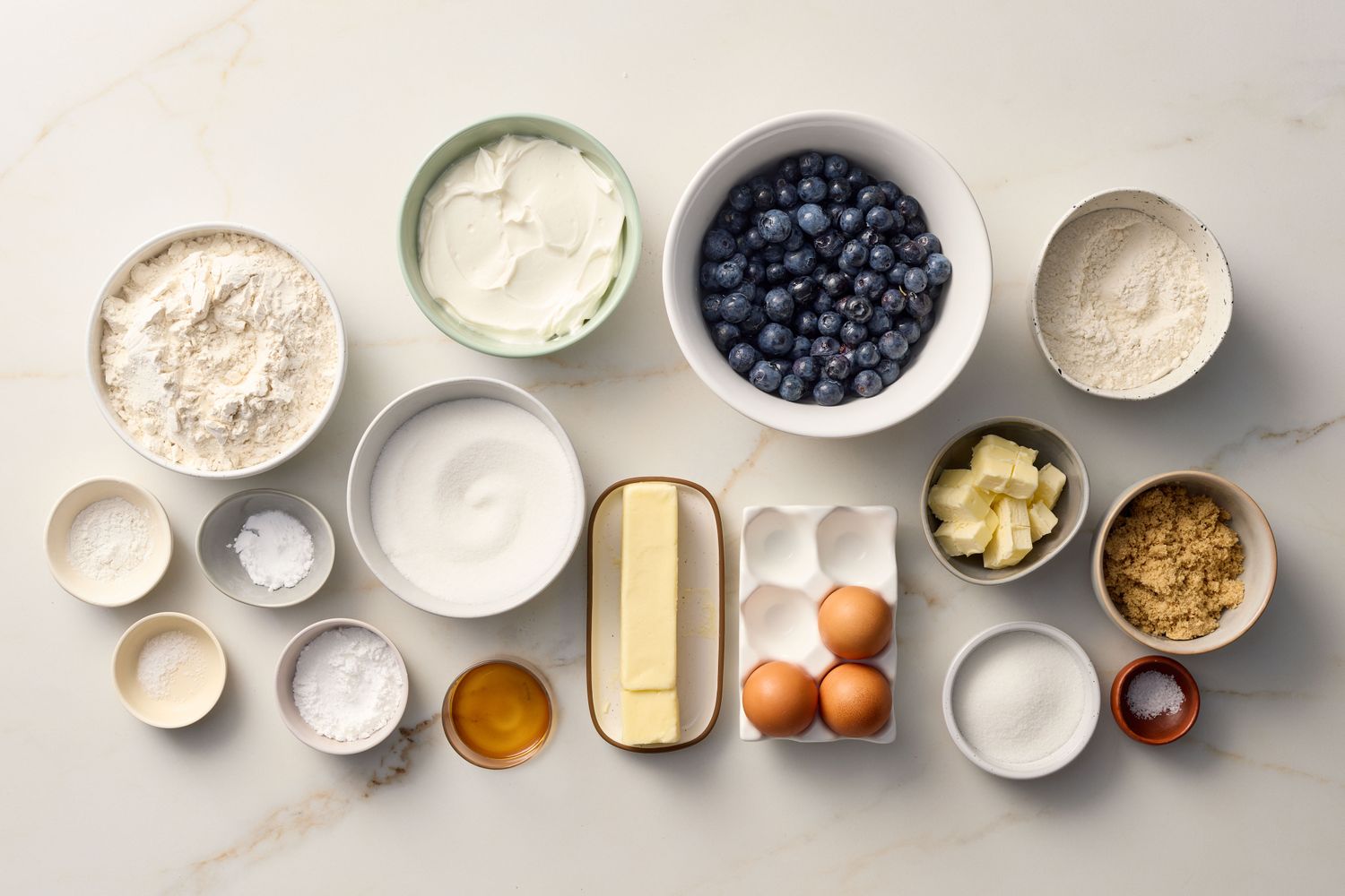 Ingredients arranged on a surface for a blueberry crumb cake recipe, including blueberries, flour, sugar, eggs, and butter
